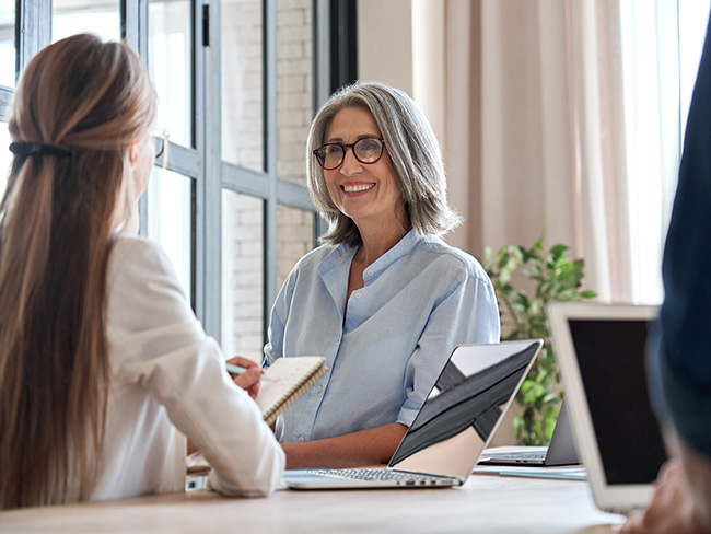 A woman in glasses gives advice to a younger woman across from her, who is taking notes on a notepad